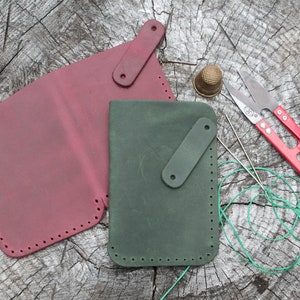 May include: Two leather wallets, one burgundy and one dark green, are laid out on a wooden surface. The wallets are stitched with holes around the edges. A pair of small red scissors, a thimble, and two needles are also on the surface.