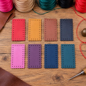 Puede incluir: Ocho parches rectangulares de cuero con bordes festoneados y agujeros preperforados en el perímetro. Los parches son de color rojo, amarillo, marrón, negro, rosa, morado, azul oscuro y verde azulado. Se ven bobinas de hilo y un punzón.