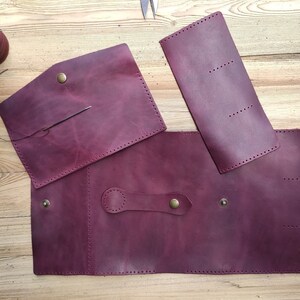 May include: Burgundy leather pieces for a wallet or purse project. The pieces are laid out on a wooden surface. The pieces have stitching around the edges and a brass button.