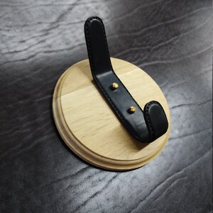 May include: A round wooden wall mount with a black leather hook. The hook is attached to the wood with two gold screws.