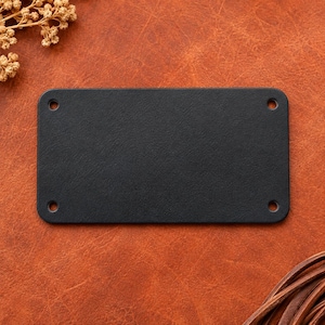 May include: A rectangular black leather patch with rounded corners and four pre-punched circular holes, one located at each corner. The patch features a smooth, matte surface texture and is positioned flat, showcasing its clean, simple design.