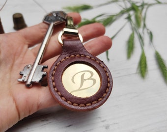 Op de afbeelding: Een bruine leren sleutelhanger met een gouden cirkel waarop de letter "B" is gegraveerd.