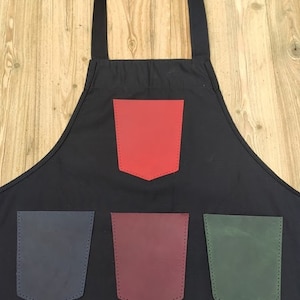 Puede incluir: Delantal negro con cuatro bolsillos de cuero. El bolsillo superior es rojo, el bolsillo inferior izquierdo es azul marino, el bolsillo inferior central es burdeos y el bolsillo inferior derecho es verde.