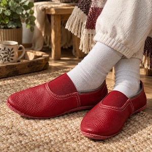 May include: A pair of red leather slip-on shoes with a textured grain finish, rounded toes, and a low-profile design. The shoes feature a flexible elasticised opening for a secure fit, visible white stitching along the seams, and a flat sole.