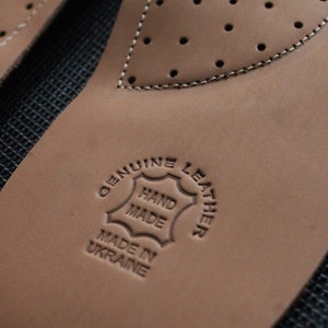 May include: Close-up of a brown leather insole with a stitched edge and a circular stamp that reads "Genuine Leather Hand Made Made in Ukraine".