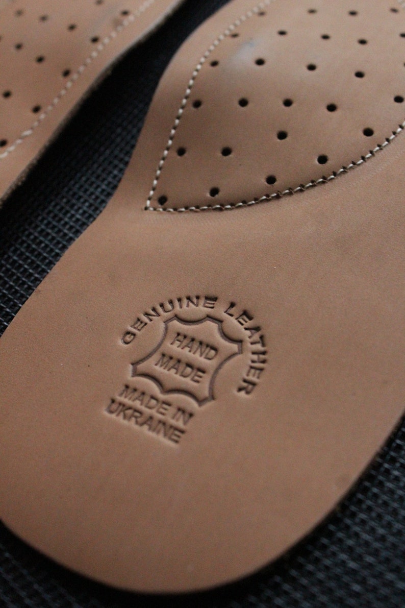 May include: Close-up of a brown leather insole with a stitched edge and a circular stamp that reads "Genuine Leather Hand Made Made in Ukraine".