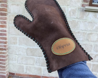 May include: Brown leather oven mitt with green stitching and an oval-shaped patch that says "Olivia" in cursive.