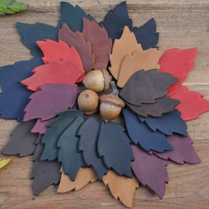 Può includere: Disposizione circolare di foglie in pelle colorate, con ghiande al centro. Le foglie sono in tonalità di rosso, marrone, blu e viola, su uno sfondo di legno. Un oggetto decorativo a tema autunnale.
