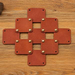 May include: A geometric arrangement of eight square, tan leather patches connected by brass-colored metal rivets. The leather squares are linked to form a cross-like pattern with open spaces between them, featuring pre-punched holes at the corners.