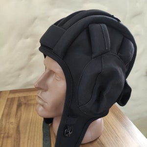 May include: Black padded helmet with a chin strap and ear flaps. The helmet is made of a soft, flexible material and is designed to protect the head from impact.