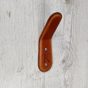 Könnte beinhalten: Ein brauner Leder-Kleiderhaken mit den Initialen "J.K." auf der Vorderseite. Der Haken ist an einer weißen Holzwand montiert.