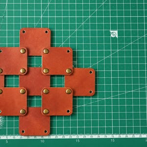 May include: A grid of seven square, rust-colored leather patches arranged in a cross pattern. Each patch features four brass-toned metal rivets at the corners, with some patches connected by these rivets. The leather has a smooth, matte finish.