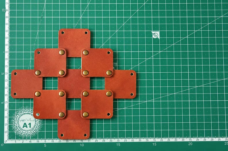 May include: A grid of seven square, rust-colored leather patches arranged in a cross pattern. Each patch features four brass-toned metal rivets at the corners, with some patches connected by these rivets. The leather has a smooth, matte finish.