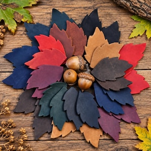 Puede incluir: Una colección de hojas de cuero troqueladas en varios colores, incluyendo rojo, azul, marrón, beige, morado y verde oscuro, dispuestas en un patrón circular. Tres bellotas marrones naturales con cúpulas texturizadas se sitúan en el centro.