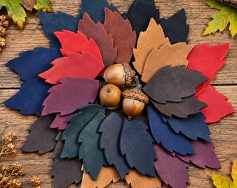 May include: A collection of die-cut leather leaves in various colours including red, blue, brown, tan, purple, and dark green, arranged in a circular pattern. Three natural brown acorns with textured caps are positioned in the centre of the leaves.