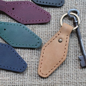 Puede incluir: Un llavero de cuero marrón con un anillo de llave de latón y una llave de estilo vintage. El llavero está cosido con un patrón de diamante y tiene un pequeño botón de latón. El llavero se muestra sobre un fondo de arpillera.
