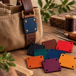 May include: A tan canvas bag with a brown leather strap and a blue leather square patch attached by four brass rivets. Several additional square leather patches in various colors including red, yellow, purple, black, and teal are scattered nearby.