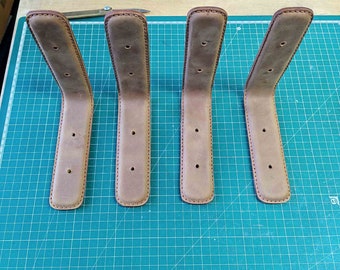 May include: Four light brown leather shelf brackets with a curved design. Each bracket has a rectangular base with three evenly spaced holes and a rounded top. The edges are stitched with a darker thread.