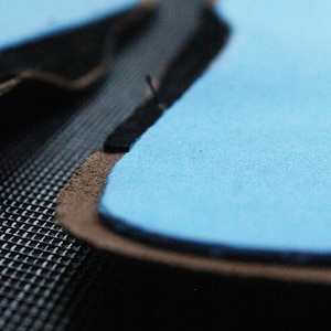 May include: Close-up of a light blue suede patch with a black stitched edge. The patch is on a dark gray textured surface.
