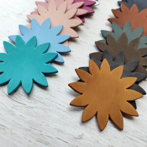 May include: Assortment of leather flower shapes in various colours, including teal, brown, blue, pink, and grey. The flowers are arranged in a row on a white wooden surface.