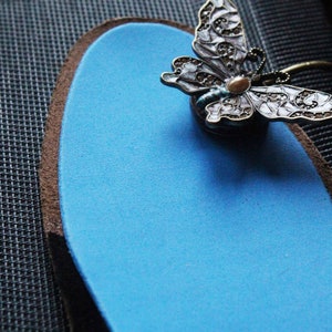 May include: A blue insole with a decorative butterfly brooch attached to it. The butterfly is made of metal and has a white and blue enamel design.