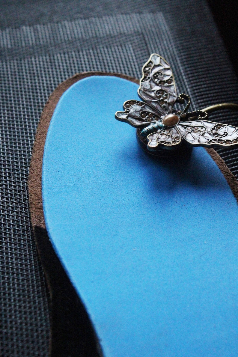 May include: A blue insole with a decorative butterfly brooch attached to it. The butterfly is made of metal and has a white and blue enamel design.