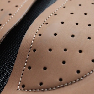 May include: Close-up of two brown leather patches with a stitched edge and a pattern of small holes. The patches are on a black background.