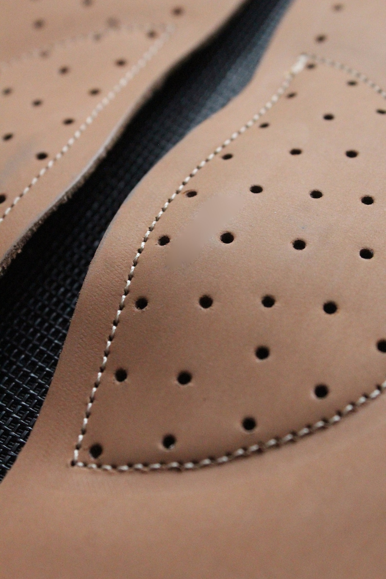 May include: Close-up of two brown leather patches with a stitched edge and a pattern of small holes. The patches are on a black background.