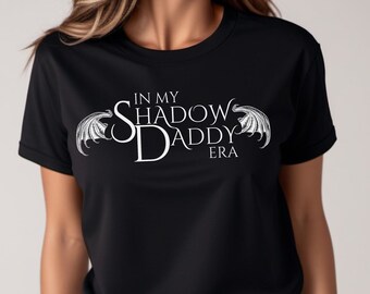 May include: Black t-shirt with the text "IN MY SHADOW DADDY ERA" in white, with decorative wing designs. The word "Black" is printed in the lower left corner. The shirt is short-sleeved.