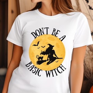 May include: White t-shirt featuring a graphic of a witch flying on a broomstick in front of a yellow moon. The text "Don't be a basic witch" is printed on the shirt.