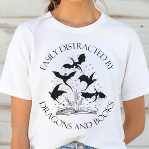 May include: White t-shirt with a black and white graphic design. The graphic features a book with dragons flying around it and the text "Easily Distracted By Dragons And Books".