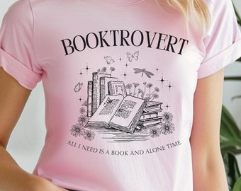 May include: Pink t-shirt with a black graphic of a stack of books, flowers, and a dragonfly. The text "BOOKTROVERT" is above the graphic and the text "ALL I NEED IS A BOOK AND ALONE TIME." is below the graphic.