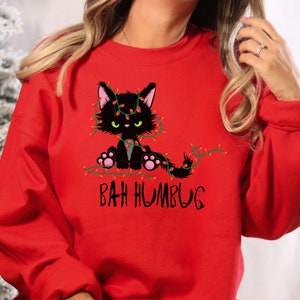 Peut inclure: Sweat-shirt rouge de Noël avec un chat noir emmêlé dans des lumières de Noël et le texte "Bah Humbug".
