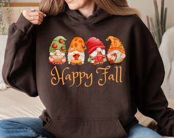 May include: Dark chocolate brown hoodie with a front graphic of four fall gnomes. Each gnome has a unique hat and holds fall-themed items. The words "Happy Fall" are written in orange script below the gnomes.