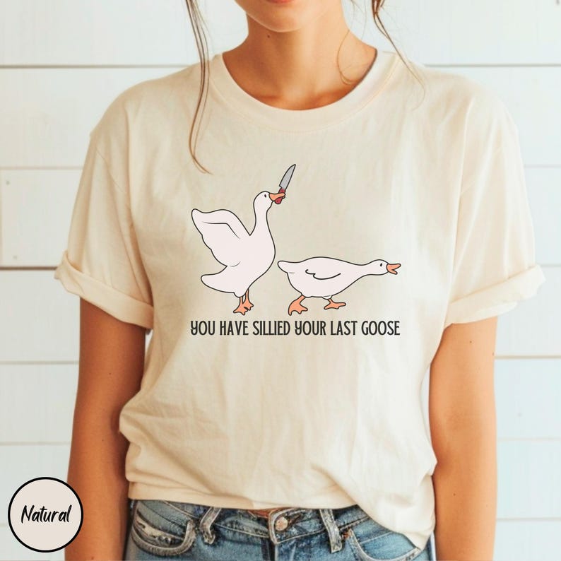 May include: A white t-shirt with a graphic of two geese. The goose on the left is holding a knife and the goose on the right is looking away. The text "You have sillied your last goose" is printed below the geese.
