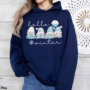 May include: A navy blue hoodie with a white snowflake and the text "hello winter" printed on the front. Four gnomes are depicted wearing blue hats and holding gifts. One gnome is holding a blue balloon.