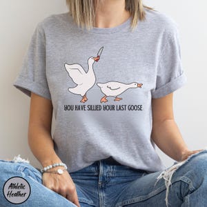 May include: A grey t-shirt with a graphic of two geese. The goose on the left is holding a knife and the text "You have sillied your last goose" is printed below the geese.