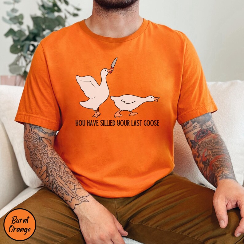 May include: A burnt orange t-shirt with a graphic of two geese. One goose is holding a knife and the other goose is running away. The text "You have sillied your last goose" is printed below the geese.