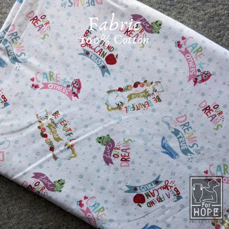 May include: White cotton fabric with a repeating pattern of whimsical illustrations and phrases. The design includes frogs, owls, apples, and text like "Live Our Dreams" and "Be Beautiful". The fabric is labeled "Fabric 100% Cotton".