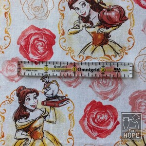May include: White fabric featuring illustrations of Belle from Beauty and the Beast, red roses, and gold decorative frames. A clear ruler with inch markings is placed over the fabric. The fabric also has the text "For HOPE Design Company".