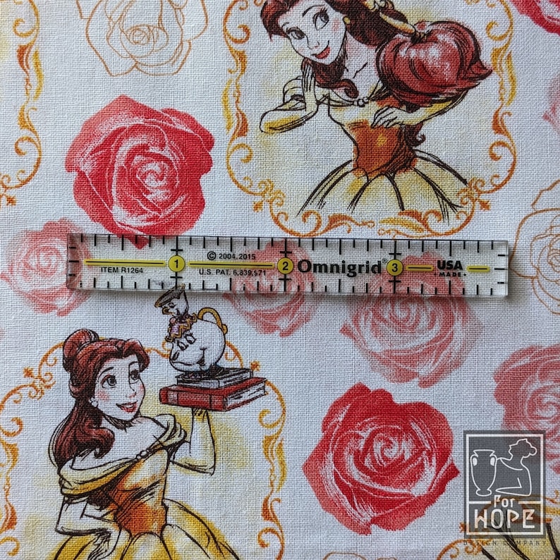 May include: White fabric featuring illustrations of Belle from Beauty and the Beast, red roses, and gold decorative frames. A clear ruler with inch markings is placed over the fabric. The fabric also has the text "For HOPE Design Company".