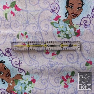 May include: Fabric featuring a repeating pattern of a cartoon character, flowers, and swirling designs in shades of purple, pink, and blue. A clear ruler with black markings and text is placed on the fabric. The ruler is marked in inches.