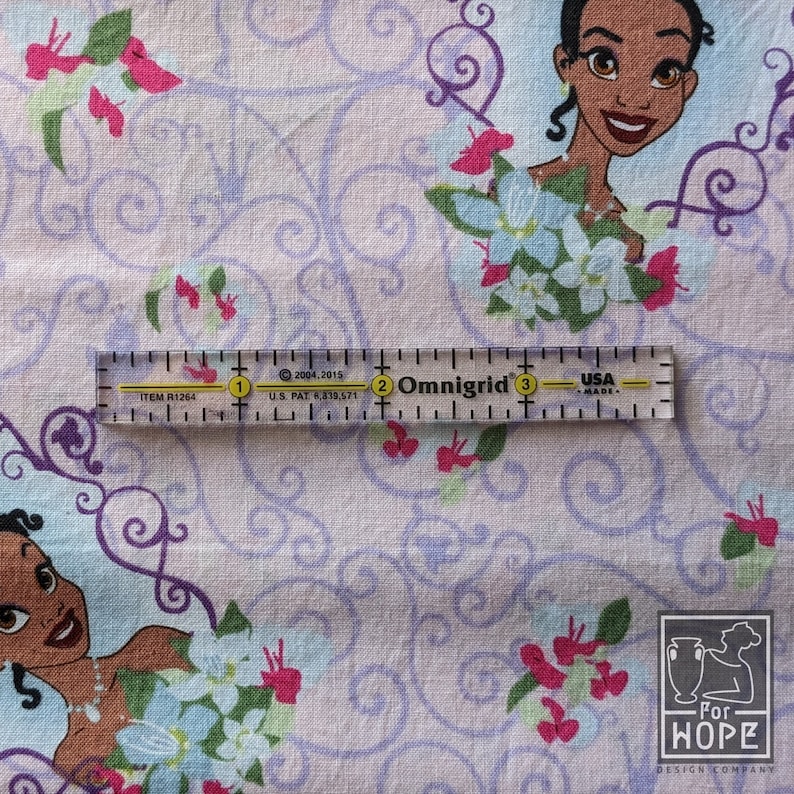 May include: Fabric featuring a repeating pattern of a cartoon character, flowers, and swirling designs in shades of purple, pink, and blue. A clear ruler with black markings and text is placed on the fabric. The ruler is marked in inches.