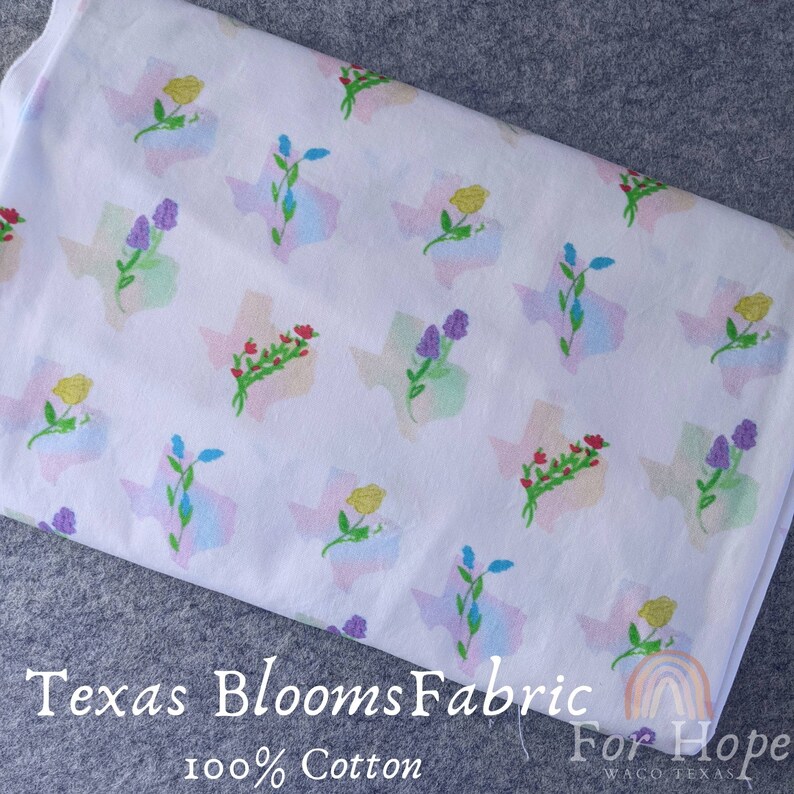 May include: White cotton fabric with a repeating pattern of the state of Texas outlined in pastel colors and a cluster of flowers in the center of each state. The text "Texas Blooms Fabric" and "100% Cotton" are printed at the bottom of the fabric.