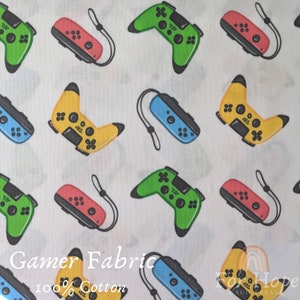 May include: White fabric with a repeating pattern of colorful video game controllers and handheld gaming devices. The fabric has the text "Gamer Fabric 100% Cotton" printed on it.
