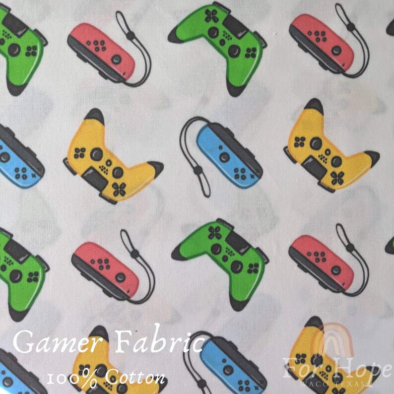 May include: White fabric with a repeating pattern of colorful video game controllers and handheld gaming devices. The fabric has the text "Gamer Fabric 100% Cotton" printed on it.
