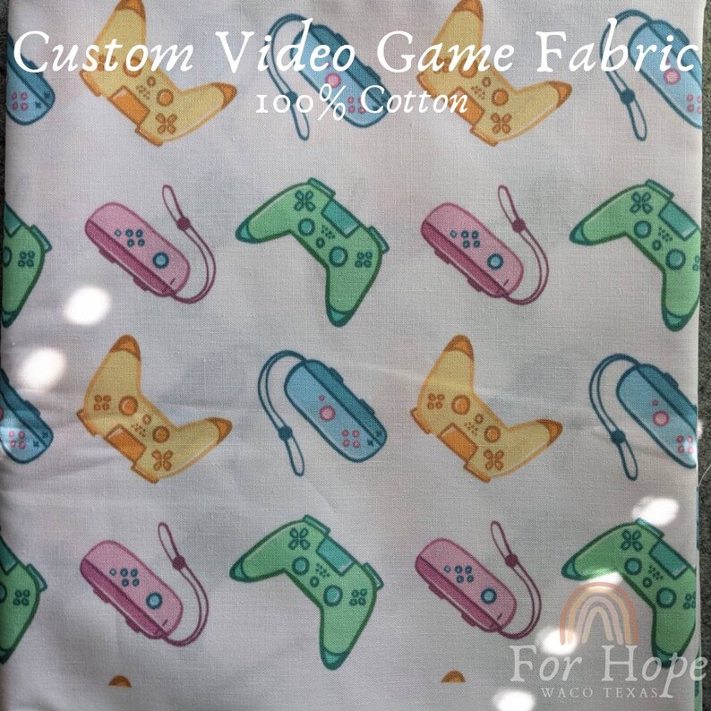 May include: White fabric with a repeating pattern of colorful video game controllers. The fabric is 100% cotton and says "Custom Video Game Fabric" and "For Hope Waco Texas" at the top and bottom.