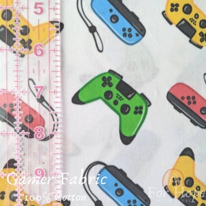 May include: White fabric with a repeating pattern of colorful video game controllers. The controllers are in various colors, including green, blue, red, and yellow. The fabric has the text "Gamer Fabric 100% Cotton" printed on it.