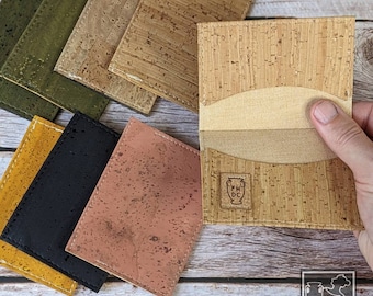 May include: A set of cork wallets in various colors, including green, brown, black, yellow, and pink. The wallets are made of natural cork and have a stitched edge. The wallets are laid out on a wooden surface. The text "For Hope Design Company" is visible in the lower right corner of the image.