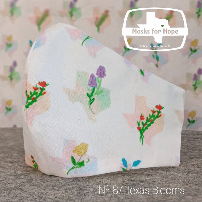 May include: White face mask with a repeating pattern of colorful Texas state outlines with floral bouquets. The text "Masks for Hope" is in a white rectangle with a red heart in the shape of Texas. The text "No. 87 Texas Blooms" is at the bottom of the image.